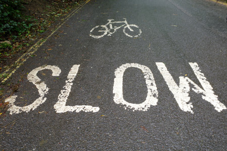 White lettering on asphalt reads 'SLOW' with a bicycle symbol above it, indicating a road combined with a cycling path. The markings are worn. Photo is taken near a village in the Cotswolds, Englandの写真素材