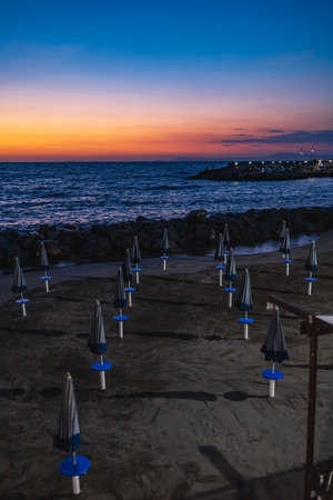 Beautiful sunset over the sea and small beach full of blue and white umbrellas next to giant rocksの写真素材