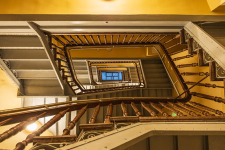 View to top of highlighted high and old square spiral staircase with decorative wooden railings and yellow ceilings with glass skylight on topの写真素材
