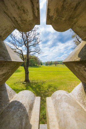 Beautiful landscape of big green park with green glades, trees at cloudy sunny morning seen through decorative hole in concrete fence of small bridge in parkの写真素材