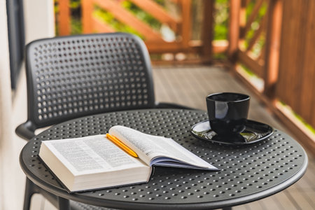 Open book with yellow pen lying on plastic garden table with black coffee cup standing on balcony...