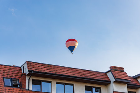 Colorful hot air balloon flying in a clear blue sky above red-tiled rooftops of residential buildings during golden hourの写真素材