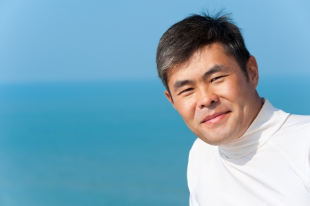 handsome asian young man looks into camera smiling, blue sea and skyの写真素材