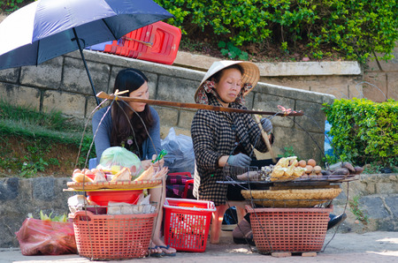 DALAT, VIETNAM - JUNE 8, 2014: An unidentified Vietnamese woman grills and sells rice pancakes, corns, sweet potatos and eggs. A young woman beside waits for her order.のeditorial素材