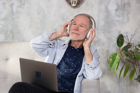 Senior man seated on sofa in living room hold laptop on lap wearing headphones listens favourite track.の写真素材