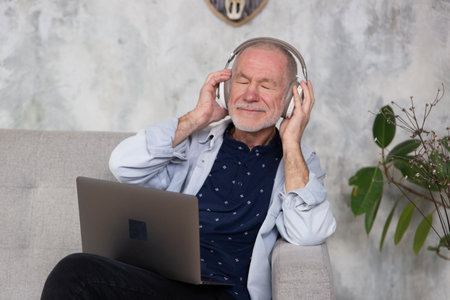 Senior man seated on sofa in living room hold laptop on lap wearing headphones listens favourite...