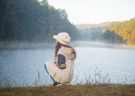 Tranquil young woman sitting on the lake, Young woman sitting outdoor Travel Lifestyle and melancholy emotions concept winter foggy nature on backgroundの写真素材