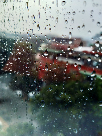 Dark clouds loom over city houses seen through a rain-streaked window. A closeup view of water...