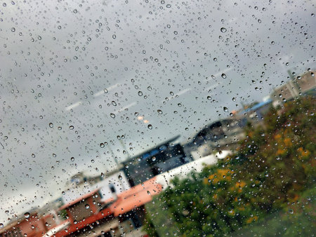 Raindrops cling to a window with city houses blurred in the background and dark clouds. A closeup...