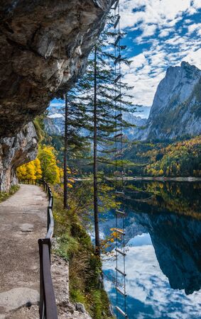 Autumn alpine view. Peaceful mountain lake with clear transparent water and reflections and ferrata stairs. Gosauseen or Vorderer Gosausee lake, Upper Austria. Dachstein summit and glacier in far.の写真素材
