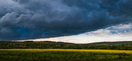 Dramatic thunderstorm cloudy sky over spring yellow flowering rapeseed fields and countryside farmlands hills. Natural weather, climate, countryside beauty concept scene.の写真素材