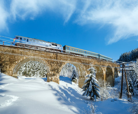Stone viaduct (arch bridge) on railway through mountain snowy fir forest. Snow drifts  on wayside and hoarfrost on trees and electric line wires.の写真素材