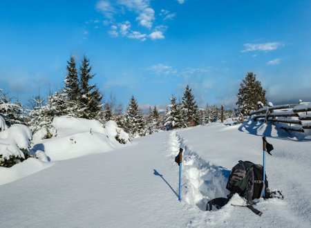 Winter remote alpine village outskirts, snow drifts on mountain fir forest edge. Tourist backpack on a freshly trodden hiking trail path.の写真素材