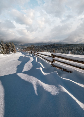 Picturesque waved shadows on snow from wood fence. Alpine mountain winter hamlet outskirts, snowy path, fir forest. High resolution image with great depth of field.の写真素材