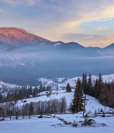 Sunrise morning winter mountain village outskirts in Black Cheremosh river valley between alp. View from rural snow covered path on hill slope with christianity crosses, Zelene, Verkhovyna, Ukraine.の写真素材
