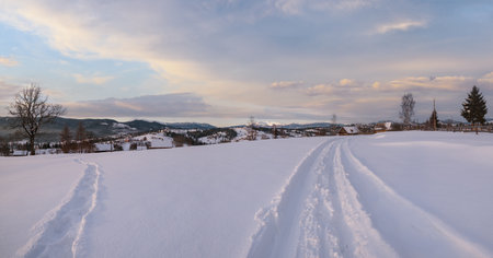 Small alpine village and winter snowy mountains in first sunrise sunlight around, Voronenko, Carpathian, Ukraine. Trace on freshly trodden path through snow drifts.の写真素材
