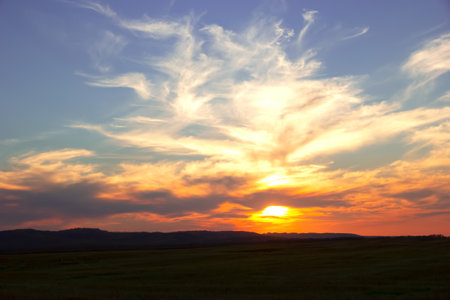 Feather clouds above the prairies and hills in the orange sunset time. Sun is behind clouds.の写真素材