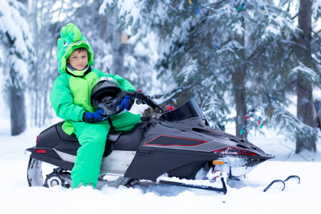 Country lifestile: caucasian boy sists in a green dragon suit on his snowmobile with helmet in hands ready for a ride.の写真素材