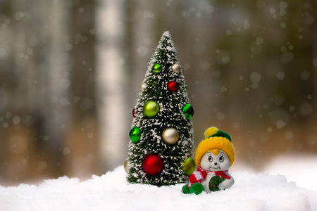 Miniature toy decorated New year tree and a little cute ceramic snowman with a yellow knitted hat in the winter forest on the snow.の写真素材