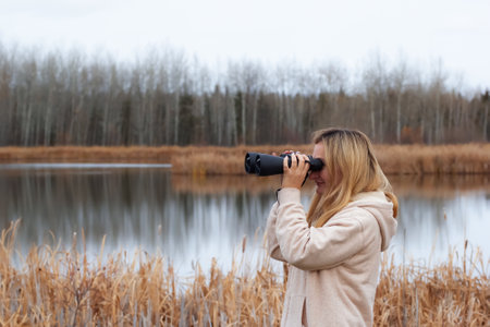 A blond caucasian woman in beige coat is watching through binoculars in the park near the lake in late autumn.の写真素材