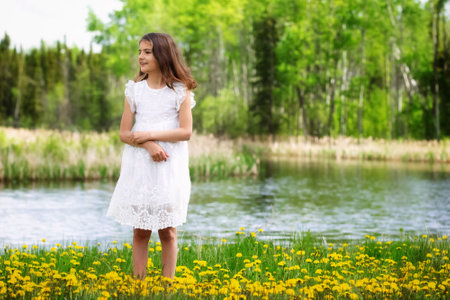 Girl in white dress is standing in green grass with blooming dandelions in the meadow in the forest near the lake.の写真素材