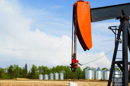 View to the oil pump in the agricultural field and row of steel grain bins along the forest in the background. Blue cloudy sky.の写真素材