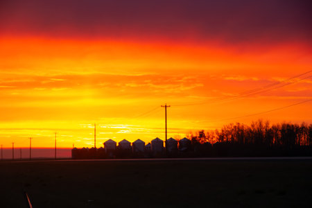 Bright beautiful sunrise with orange and purple light along the highway and silhouettes of a grain bins at the farm in the horizon in autumn.の写真素材