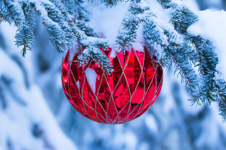 Large red Christmas ball of geometric pattern is hanging on the spruce tree branch covered with snow.の写真素材