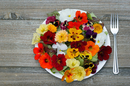 Beautiful healthy vegan salad of lettuce and colourful edible  flowers (nasturtium, calendula and pansy) on the wooden garden table with a fork on a side.の写真素材