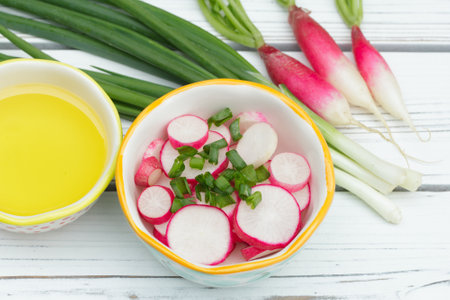 Salad of sliced raddishes (French breakfast variety) and green onions in a little bowl, all ingredients with olive oil are on the white wooden rustic table.の写真素材