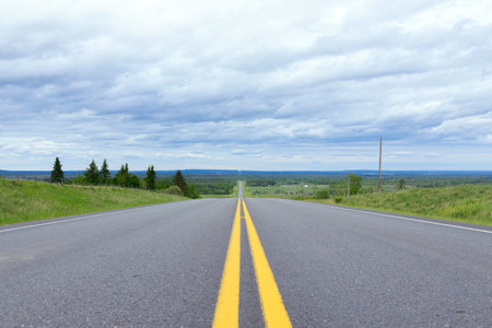 Highway with double solid yellow lines in prairies with green grass and trees along, blue cloudy sky...