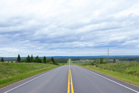 Highway with double solid yellow lines in prairies with green grass and trees along, blue cloudy sky above.の写真素材