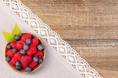 Wooden plate with vibrant summer berries, blueberries and raspberries with green leaves, on a gray cotton table cloth with white laceの写真素材