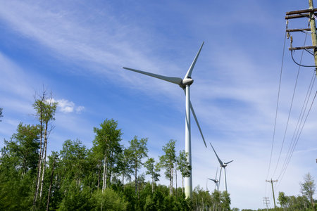 Row of wind turbines producing sustainable energy, power line along in the woodland and blue sky...