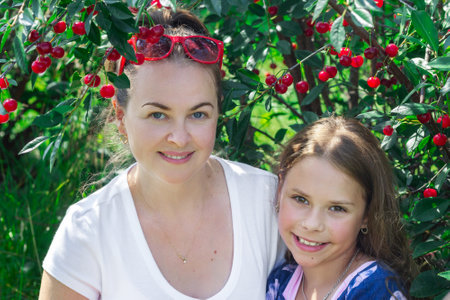 Mother and daughter are hugging and smiling together in a summer orchard among branches with ripe...