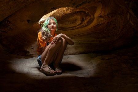 A teenager girl with green hair is sitting alone in darkness in a wonderful cave with spiral pattern and light beam from the ceiling. Gundy caves, British Columbia, Canada.の写真素材