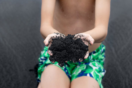 A teen boy in vivid swimming trunks is sitting in the beach and holding a handful of unique...