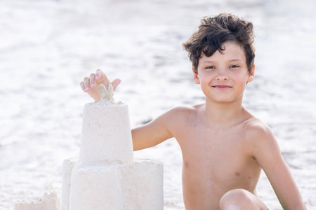 White tanned teenager boy is sitting on a white sand at the ocean shore and building a sandcastle on summer vacations.の写真素材