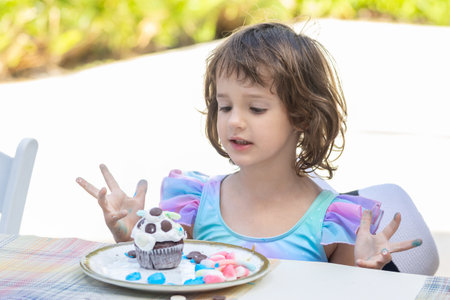 Cute little toddler girl in bright blue and pink dress is sitting at the table outdoors and...