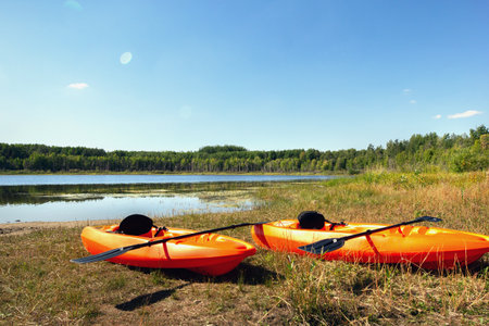 Couple of bright orange kayaks are at the shore of the little country lake amidst forest in sunny summer day. Blue sky and peaceful nature.の写真素材