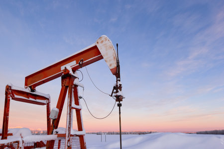 Oil pumpjack covered in snow against a soft pink winter sky at sunrise. Industrial energy production in rural Canada, cold season, oil and gas industry, prairie landscape.の写真素材
