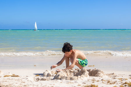 Teenager boy is playing with sand at the tropical beach, turquoise waters of the ocean are on the background, hot sunny day at the shore.の写真素材