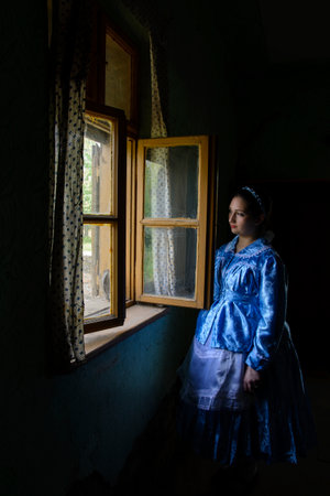 Ivanovo, Vojvodina, Serbia â april 17, 2016: Girl dressed in a traditional Paulician clothing is standing next to an open window in an old house.のeditorial素材
