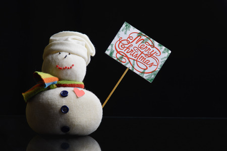 Snowman figure holding a paperboard sign on a stick with text âMerry Christmas!â written on it, and surrounded with fir branches. Contrasty low key photograph.の写真素材