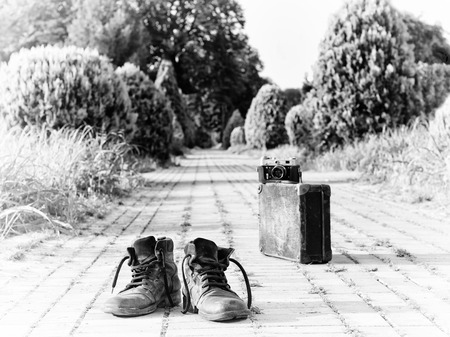 Old ankle leather boots in a foreground, in the middle of a brick road. A film camera on an antique cardboard suitcase with corner protectors, in a blurry background. A black-and-white photo effect.の写真素材