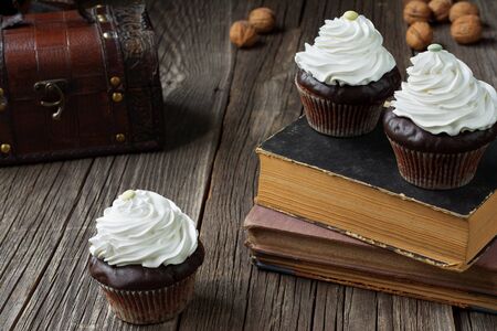 Cupcakes in paper patty cases, topped with whipped cream, are lying on antique books and rustic weathered wood planks. A chest and a few walnut shells are visible in a blurry background.の写真素材