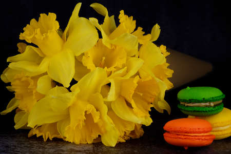 A bouquet of Daffodils in a paper cone and three colorful macarons (green, orange, yellow) are lying...