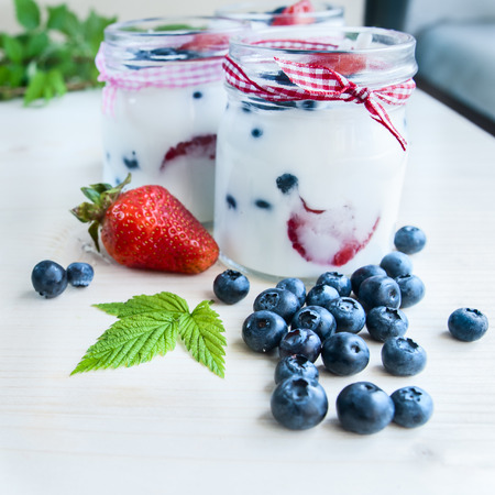 Three jars of yogurt with blueberries and strawberries, square. Berries of blueberries and strawberries are scattered on the table. Cooking, healthy food, food packagingの写真素材