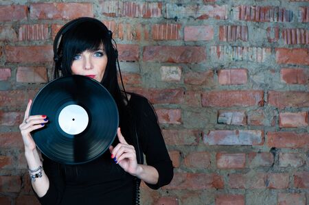 Trendy beautiful girl brunette DJ with a vinyl record in her hands, looking into the camera, against a background of a brick wall, space for text. Theme of musicの写真素材