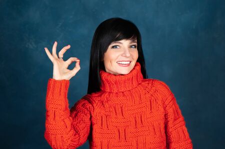 Trendy beautiful girl, brunette in a bright orange sweater, shows an ok sign and smiling . The theme of autumn and coolness. Studio portrait of a woman on a monochrome blue, trendy shabby backgroundの写真素材